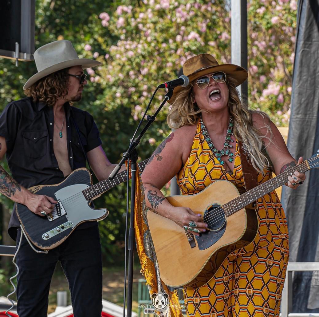 Woman wearing yellow dress and hat playing acoustic guitar and singing into a microphone onstage next to a man in a hat playing electric guitar.